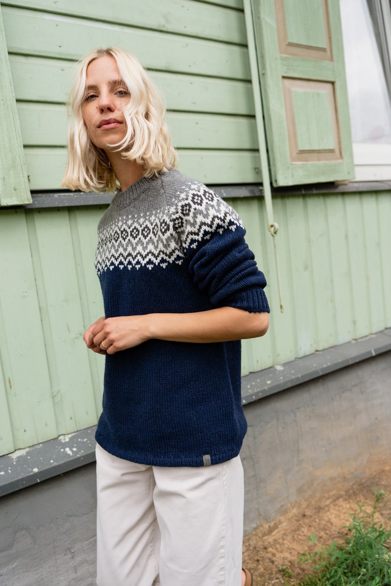 May include: A woman wearing a navy blue sweater with a grey and white geometric pattern. The sweater has a crew neck and long sleeves. She is wearing white pants.
