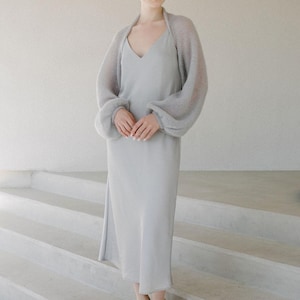 May include: A light gray midi-length slip dress with a V-neckline paired with a matching sheer, long-sleeved knit bolero shrug. The outfit is completed with light gray open-toe sandals featuring decorative crystal embellishments on the front straps.