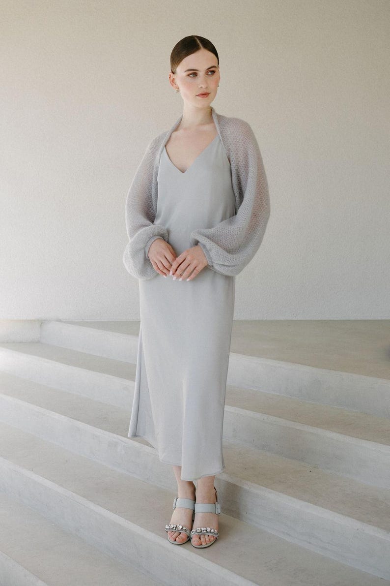 May include: A light gray midi-length slip dress with a V-neckline paired with a matching sheer, long-sleeved knit bolero shrug. The outfit is completed with light gray open-toe sandals featuring decorative crystal embellishments on the front straps.