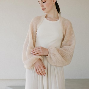 May include: A sheer, pale beige bolero shrug featuring a loose, open-knit texture and voluminous bishop sleeves with fitted cuffs. The garment is worn over a simple, sleeveless cream-colored dress with a round neckline and a pleated skirt.