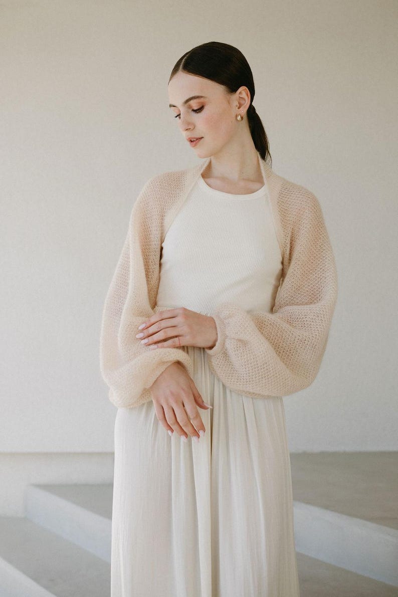 May include: A sheer, pale beige bolero shrug featuring a loose, open-knit texture and voluminous bishop sleeves with fitted cuffs. The garment is worn over a simple, sleeveless cream-colored dress with a round neckline and a pleated skirt.