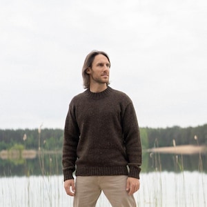Peut inclure: Une personne porte un pull ras du cou marron et un pantalon kaki près d'un lac. Le pull est de couleur marron foncé et semble être fait d'une matière texturée. L'arrière-plan présente un lac et des arbres.