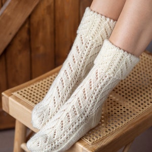 May include: A pair of white knitted socks with a lacy pattern. The socks are on a wooden stool with a woven seat.