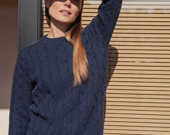 Puede incluir: Un suéter de manga larga en color azul marino oscuro con un patrón clásico de punto trenzado, cuello redondo y puños y dobladillo acanalados. La prenda presenta un diseño de punto texturizado que recorre verticalmente el frente.