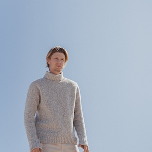 Puede incluir: Un hombre con un su&eacute;ter gris claro de cuello alto y pantalones beige se para frente a un cielo azul.