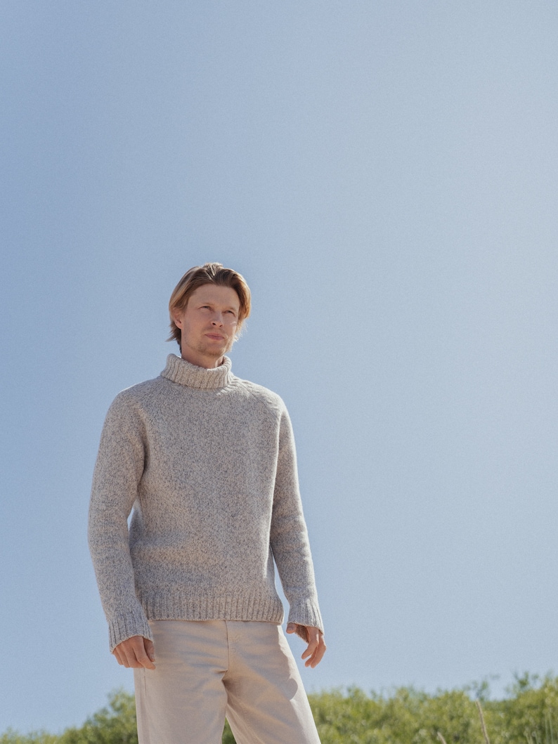 Puede incluir: Un hombre con un su&eacute;ter gris claro de cuello alto y pantalones beige se para frente a un cielo azul.