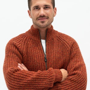 Pu&ograve; includere: Un uomo indossa un cardigan con zip in maglia color ruggine con una trama a coste. &Egrave; in piedi con le braccia incrociate e guarda direttamente la telecamera.