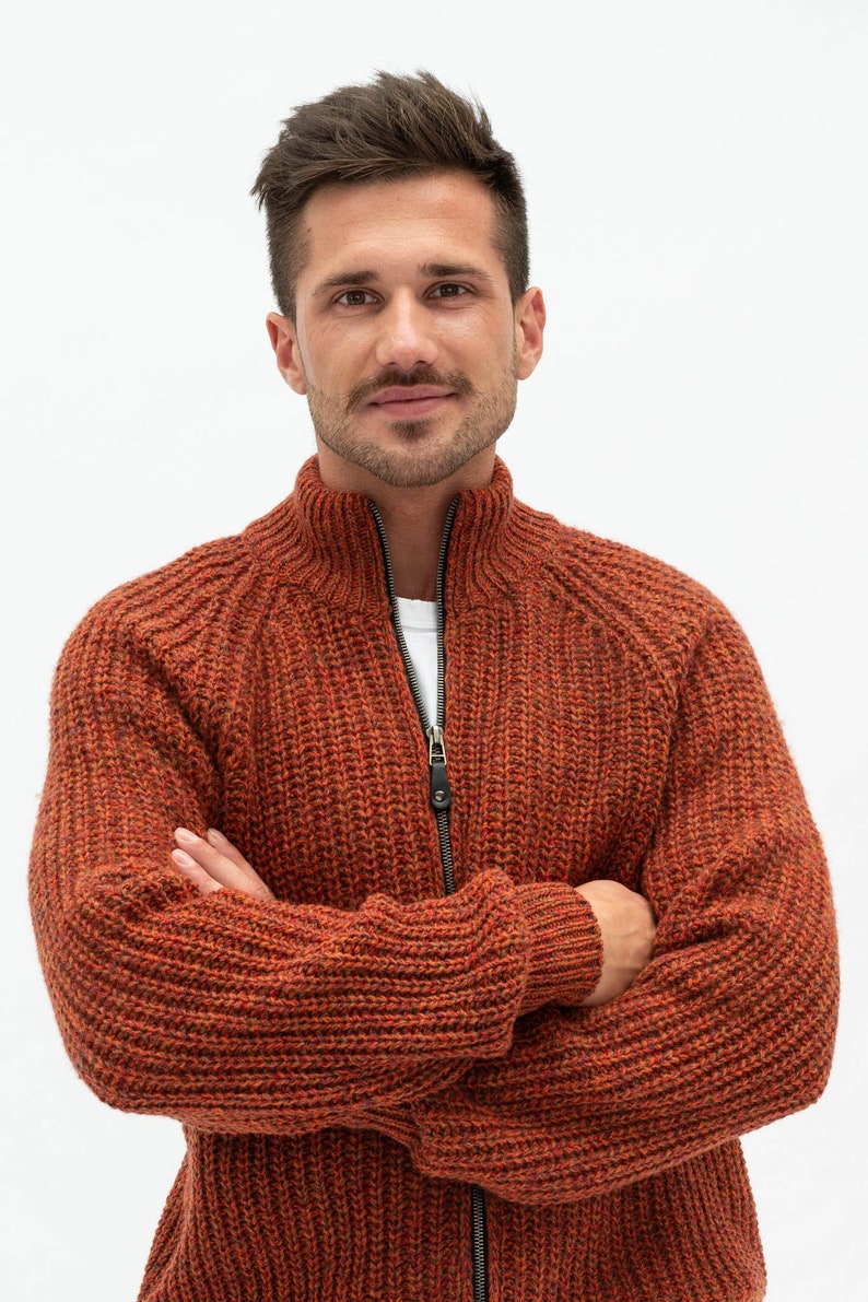Pu&ograve; includere: Un uomo indossa un cardigan con zip in maglia color ruggine con una trama a coste. &Egrave; in piedi con le braccia incrociate e guarda direttamente la telecamera.
