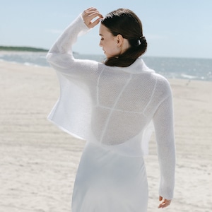 May include: A white long-sleeved cropped bolero sweater featuring a fine knit texture and an open front design, layered over a matching white sleeveless dress. The garment has a wide collar and a loose, boxy fit with long sleeves.
