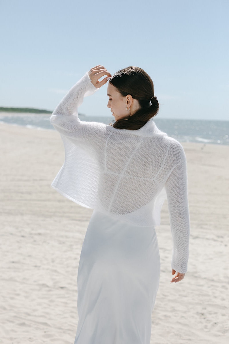 May include: A white long-sleeved cropped bolero sweater featuring a fine knit texture and an open front design, layered over a matching white sleeveless dress. The garment has a wide collar and a loose, boxy fit with long sleeves.