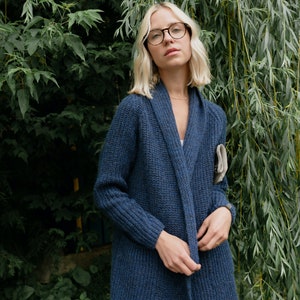 Pu&ograve; includere: Una donna con occhiali rotondi e un lungo cardigan di maglia blu scuro con collo a scialle. Il cardigan ha tasche sui lati ed &egrave; aperto sul davanti. &Egrave; in piedi davanti a uno sfondo verde frondoso.
