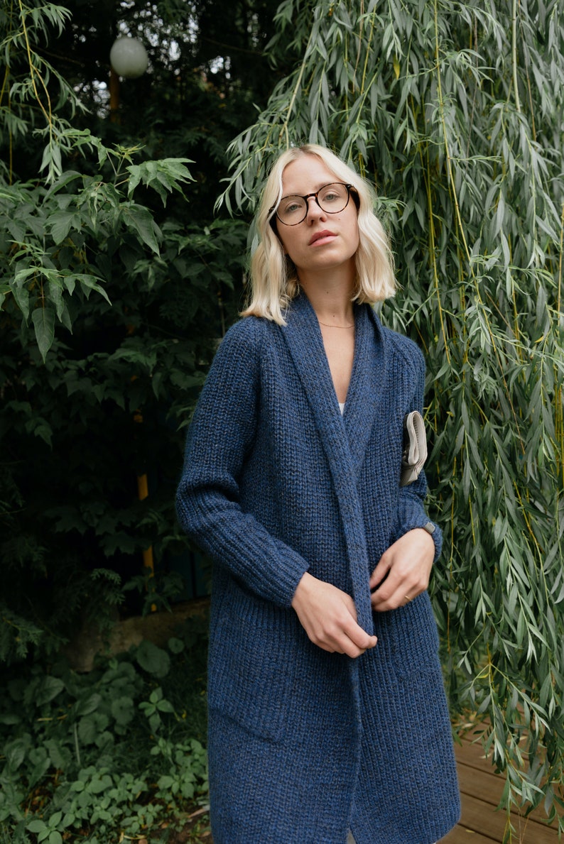 Pu&ograve; includere: Una donna con occhiali rotondi e un lungo cardigan di maglia blu scuro con collo a scialle. Il cardigan ha tasche sui lati ed &egrave; aperto sul davanti. &Egrave; in piedi davanti a uno sfondo verde frondoso.