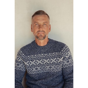 May include: A navy blue crew neck sweater featuring a white geometric design across the chest and shoulders. The sweater has long sleeves and a textured knit. The person wearing the sweater has short, grey hair and a beard.