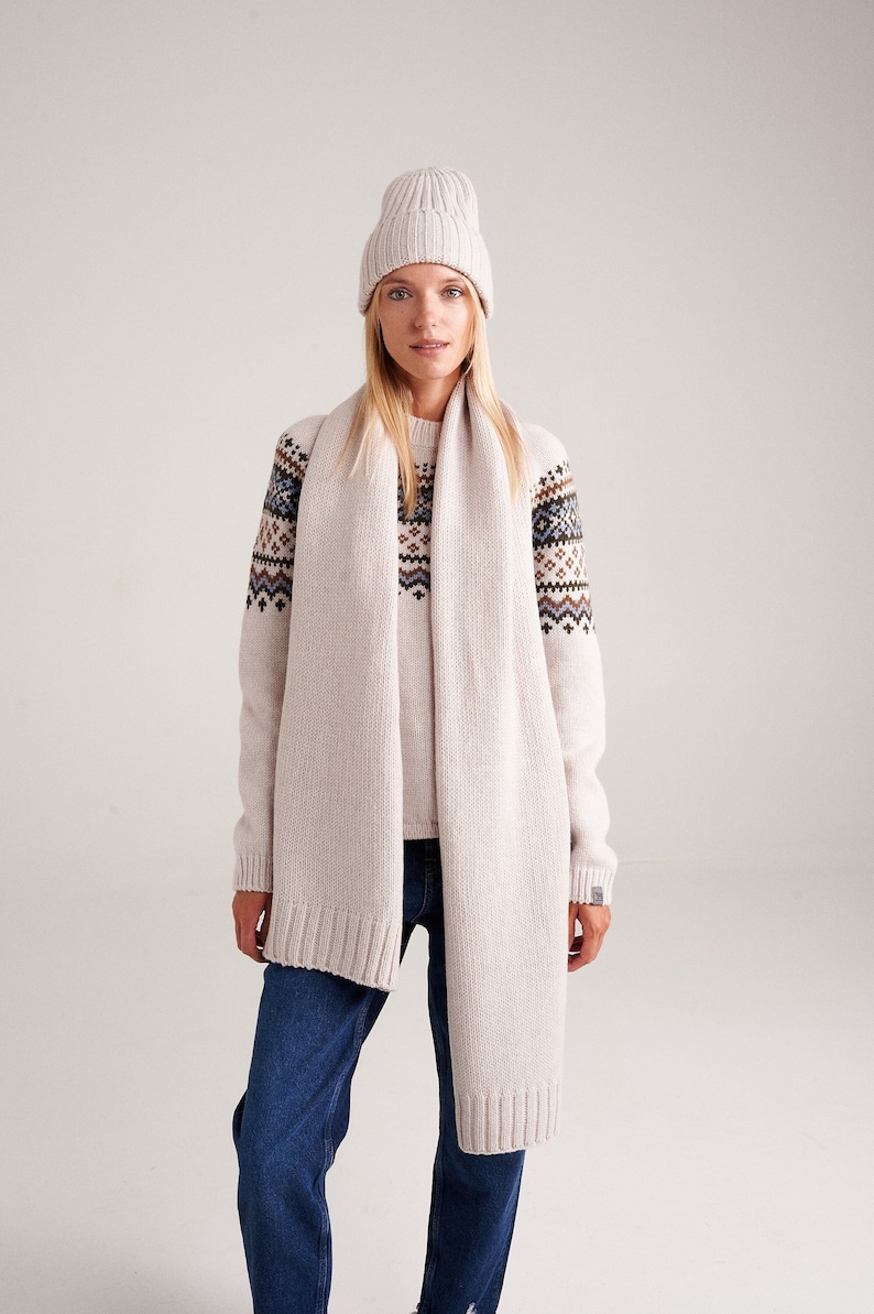 May include: A beige ribbed knit beanie, a long beige knit scarf, and a beige long-sleeve sweater featuring a brown, blue, and white geometric pattern across the chest and shoulders, paired with dark blue denim jeans.