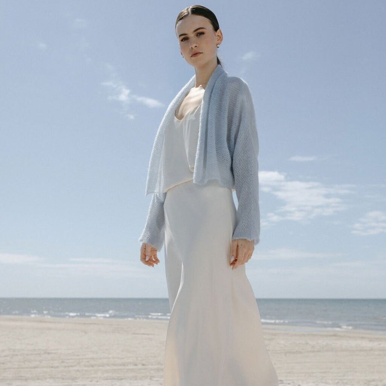 May include: A light blue open-front knit cardigan with long sleeves and a soft texture, worn over a simple white sleeveless dress. The cardigan features a relaxed fit and a draped collar, providing a lightweight layer over the smooth white dress.