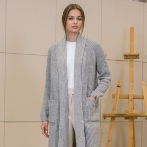 Pu&ograve; includere: Un cardigan di maglia grigia con collo a scialle e due tasche. Il cardigan &egrave; aperto e la modella indossa una t-shirt bianca sotto.