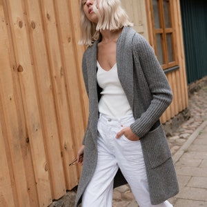 Pu&ograve; includere: Una donna indossa un cardigan grigio con cappuccio sopra una canottiera bianca e jeans bianchi. Sta camminando su un sentiero lastricato.