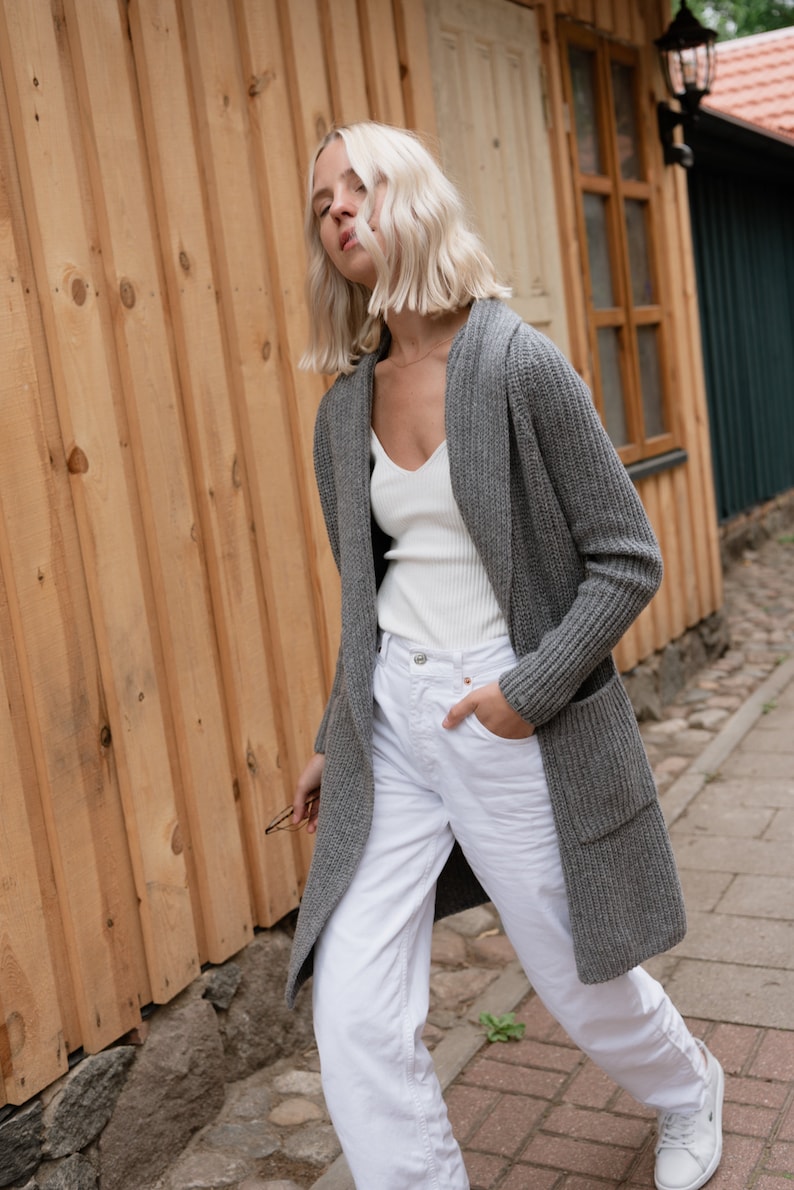 Pu&ograve; includere: Una donna indossa un cardigan grigio con cappuccio sopra una canottiera bianca e jeans bianchi. Sta camminando su un sentiero lastricato.