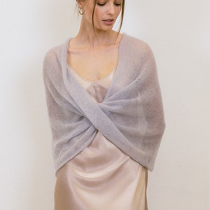 May include: A soft, light lavender-grey knitted wrap with a sheer, fuzzy texture. The wrap is styled in a crossover design across the chest, layered over a smooth, champagne-colored satin slip dress with thin straps and a V-neckline.