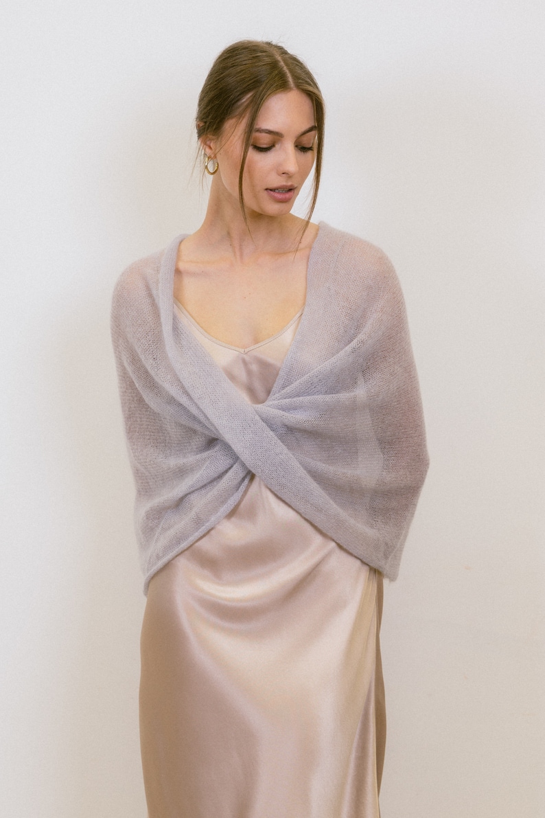 May include: A soft, light lavender-grey knitted wrap with a sheer, fuzzy texture. The wrap is styled in a crossover design across the chest, layered over a smooth, champagne-colored satin slip dress with thin straps and a V-neckline.
