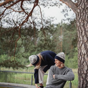 May include: Two people wearing knitted hats with a geometric pattern in blue and white. One person is wearing a navy blue sweater and white pants, while the other person is wearing a gray sweater and dark blue pants. They are sitting on a wooden bench in a forest setting.