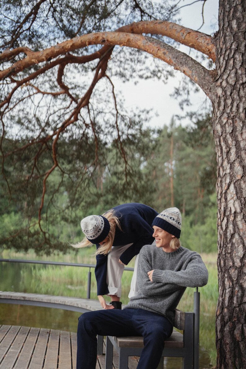 May include: Two people wearing knitted hats with a geometric pattern in blue and white. One person is wearing a navy blue sweater and white pants, while the other person is wearing a gray sweater and dark blue pants. They are sitting on a wooden bench in a forest setting.