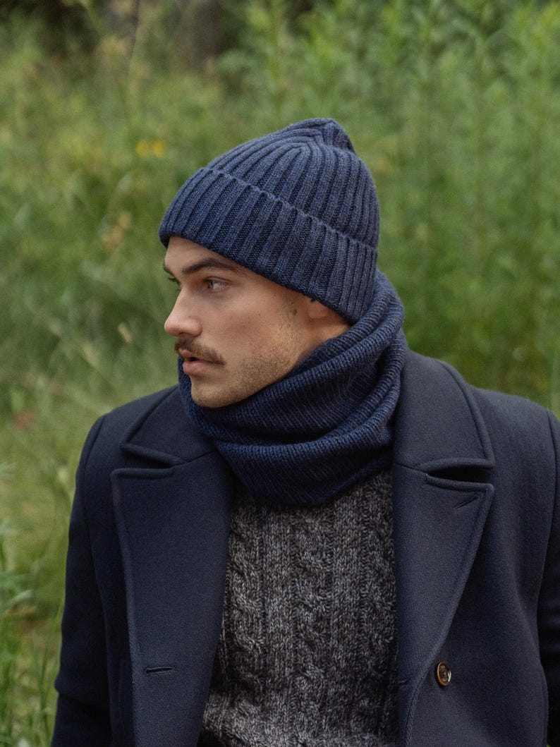May include: A dark blue ribbed knit beanie and a matching dark blue ribbed knit neck warmer. The beanie features a folded cuff design, and both items are crafted from a thick, textured yarn, providing a cohesive look when worn together.