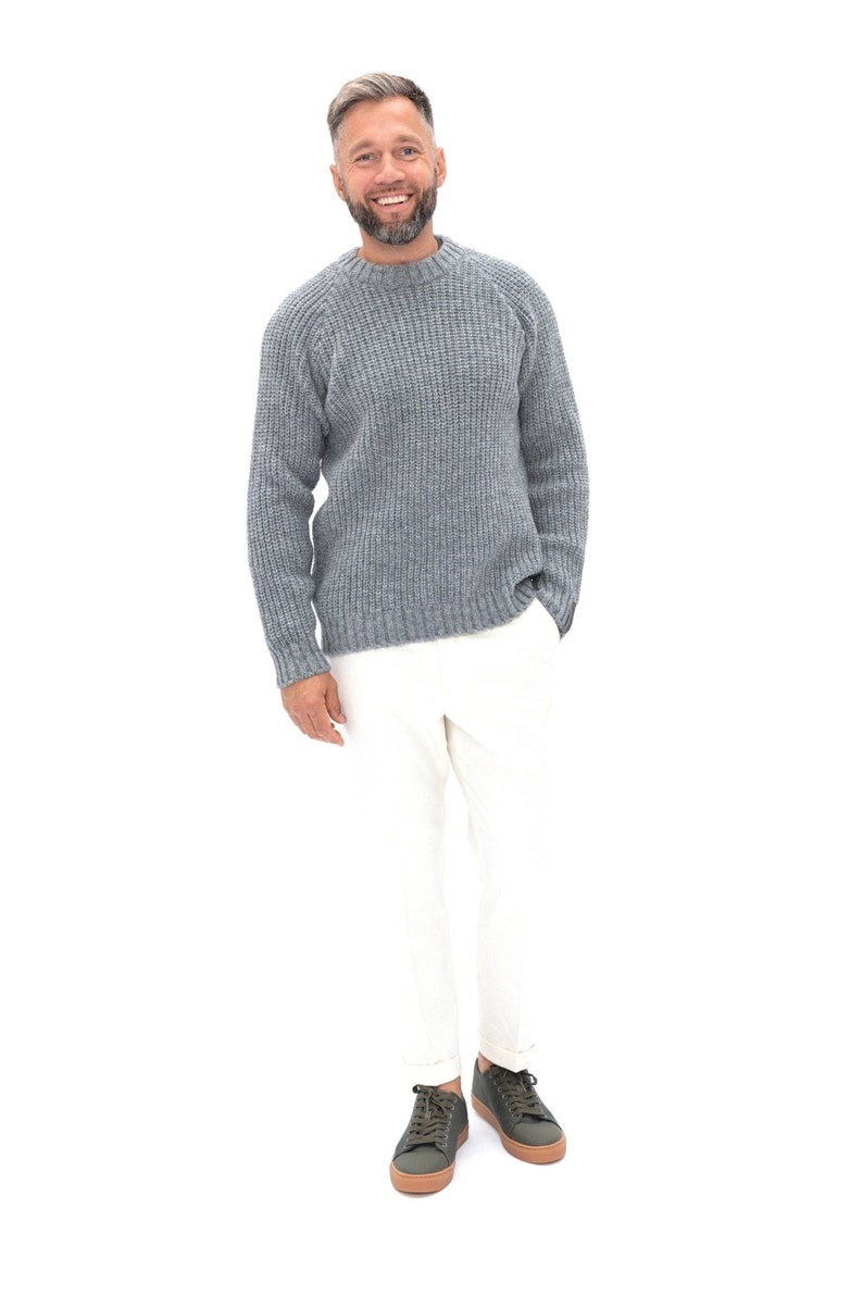May include: A man wearing a gray knitted sweater and white pants. He is smiling and looking at the camera. He is wearing green sneakers with brown soles.