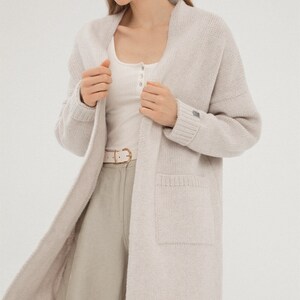 May include: A long, light beige cardigan with a shawl collar and a patch pocket on the left side. The cardigan is made of a soft, knitted fabric.