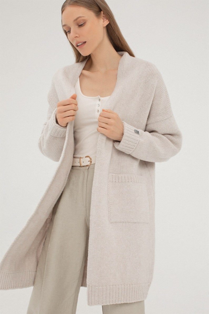 May include: A long, light beige cardigan with a shawl collar and a patch pocket on the left side. The cardigan is made of a soft, knitted fabric.
