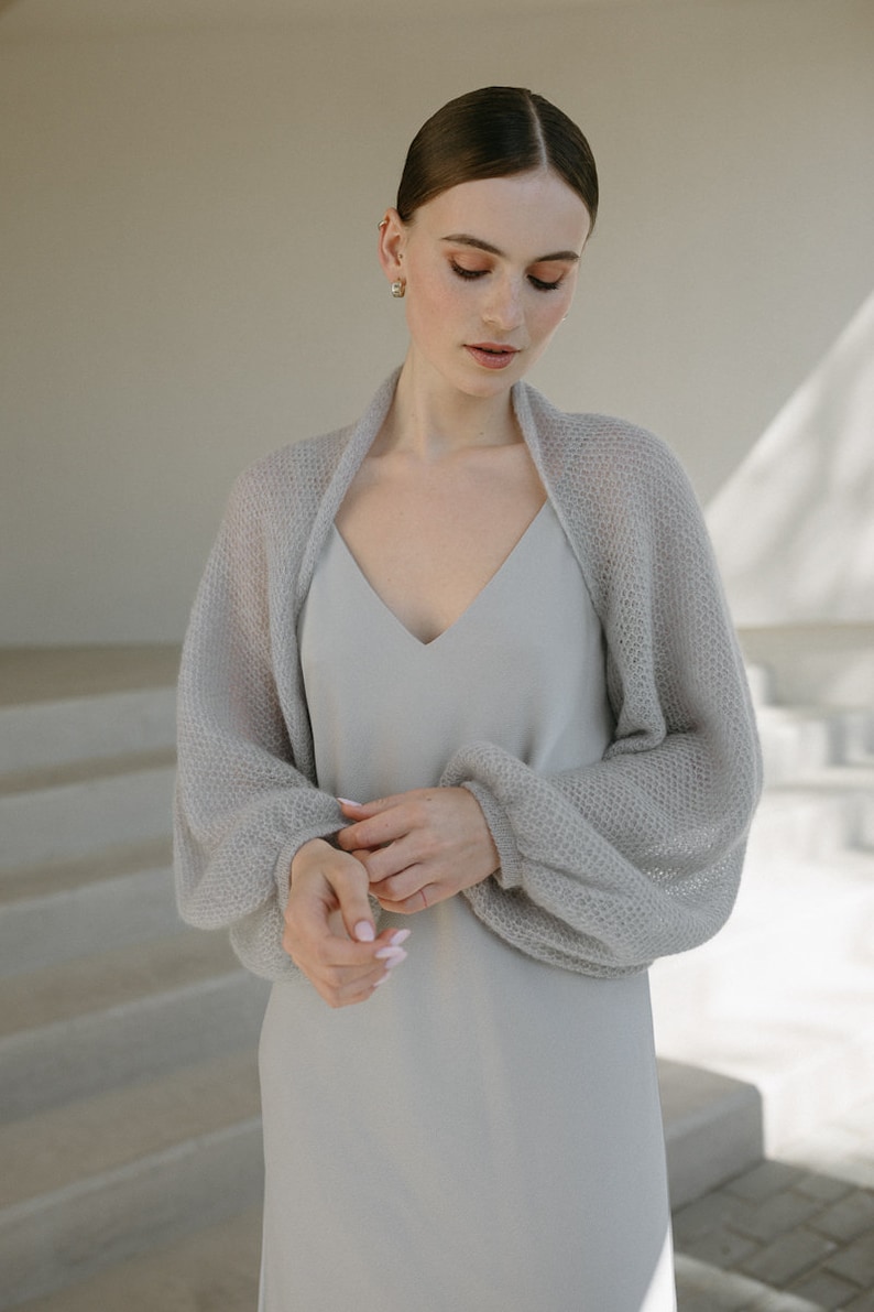 May include: A light gray sleeveless dress with a V-neckline paired with a matching light gray sheer knit bolero. The bolero features long, voluminous balloon sleeves with fitted cuffs, crafted from a delicate, open-weave, semi-transparent yarn.