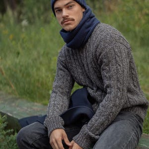 May include: A dark blue ribbed knit beanie and a matching dark blue neck warmer worn with a grey cable knit sweater and dark grey jeans. The sweater features a textured cable pattern throughout the sleeves and body, paired with a solid dark blue scarf.