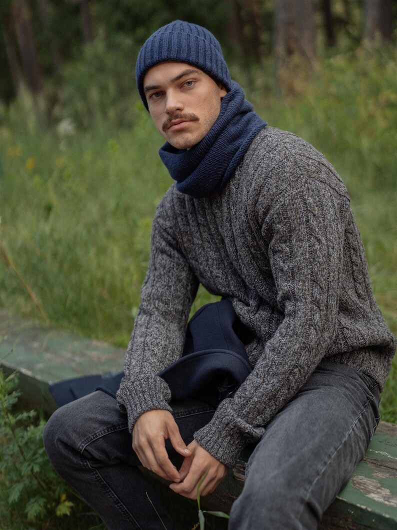May include: A dark blue ribbed knit beanie and a matching dark blue neck warmer worn with a grey cable knit sweater and dark grey jeans. The sweater features a textured cable pattern throughout the sleeves and body, paired with a solid dark blue scarf.