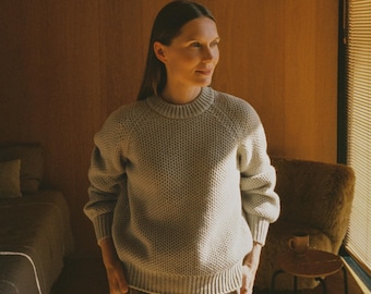 May include: A light gray, long-sleeved crewneck sweater with a textured, honeycomb-like knit pattern. The garment features ribbed cuffs, a ribbed hem, and a ribbed neckline, paired with tan, wide-leg trousers with visible front pleats.