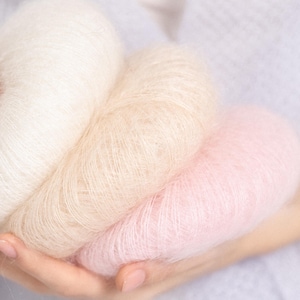 May include: Three soft, fuzzy balls of yarn in a gradient of colors, including a bright white, a warm beige, and a pale pastel pink. The yarn has a distinct halo texture, appearing light and airy with fine, delicate fibers visible throughout.
