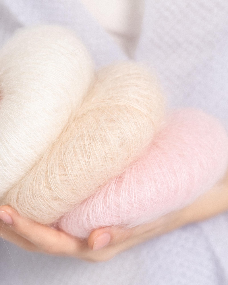 May include: Three soft, fuzzy balls of yarn in a gradient of colors, including a bright white, a warm beige, and a pale pastel pink. The yarn has a distinct halo texture, appearing light and airy with fine, delicate fibers visible throughout.