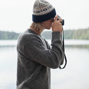 May include: A person wearing a gray sweater and a patterned beanie is taking a photo with a vintage camera. The person is standing in front of a body of water.