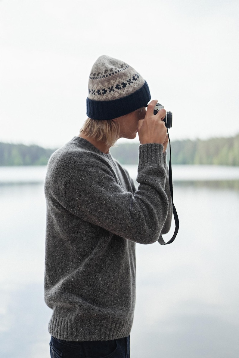 May include: A person wearing a gray sweater and a patterned beanie is taking a photo with a vintage camera. The person is standing in front of a body of water.