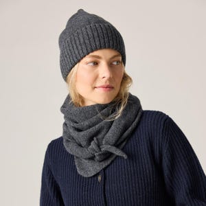 May include: A dark gray knit beanie with a ribbed cuff and a matching dark gray knit scarf tied in a knot at the front. The beanie and scarf are made of a textured wool-like material, providing a cohesive look for cold weather accessories.