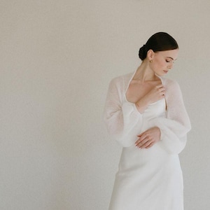 May include: A white sheer bolero with long, voluminous puff sleeves and a soft, fuzzy texture, worn over a matching white sleeveless dress with a scoop neckline. The garment features a delicate, lightweight knit construction.