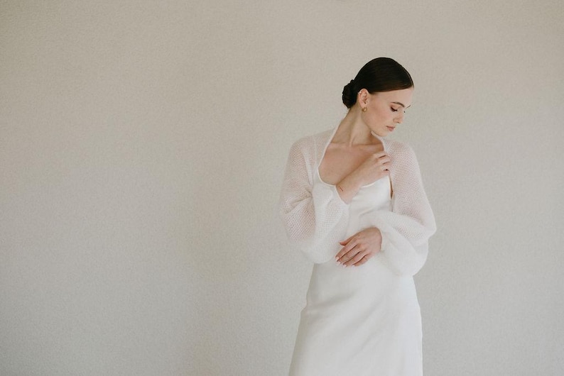 May include: A white sheer bolero with long, voluminous puff sleeves and a soft, fuzzy texture, worn over a matching white sleeveless dress with a scoop neckline. The garment features a delicate, lightweight knit construction.