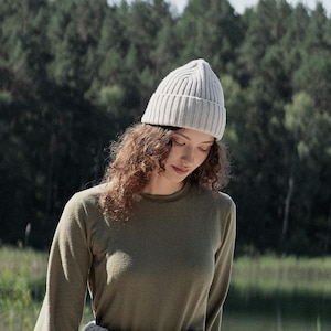 May include: A light beige ribbed knit beanie worn with a long-sleeved olive green crew neck shirt and a cream-colored cable knit sweater tied around the waist, featuring a textured knit pattern and a folded brim on the beanie.