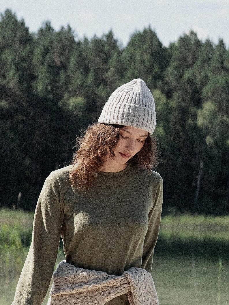 May include: A light beige ribbed knit beanie worn with a long-sleeved olive green crew neck shirt and a cream-colored cable knit sweater tied around the waist, featuring a textured knit pattern and a folded brim on the beanie.