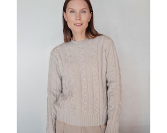 May include: A light beige cable-knit sweater with a crew neck. The sweater has a textured pattern and long sleeves. The model is wearing the sweater with beige pants. The sweater is made of a soft, comfortable material.