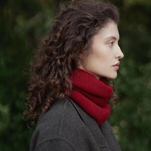 May include: A deep red knitted cowl neck scarf worn around the neck, featuring a ribbed texture and a thick, folded design. The scarf is paired with a dark charcoal gray wool coat, creating a contrast between the vibrant red and neutral gray.