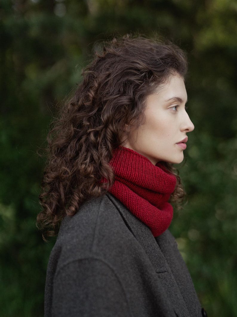 May include: A deep red knitted cowl neck scarf worn around the neck, featuring a ribbed texture and a thick, folded design. The scarf is paired with a dark charcoal gray wool coat, creating a contrast between the vibrant red and neutral gray.
