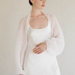 May include: A white knitted bolero with long, voluminous sleeves and a soft, textured finish, worn over a simple white slip dress with a scoop neckline. The garment features a lightweight, semi-sheer knit construction with ribbed cuffs.