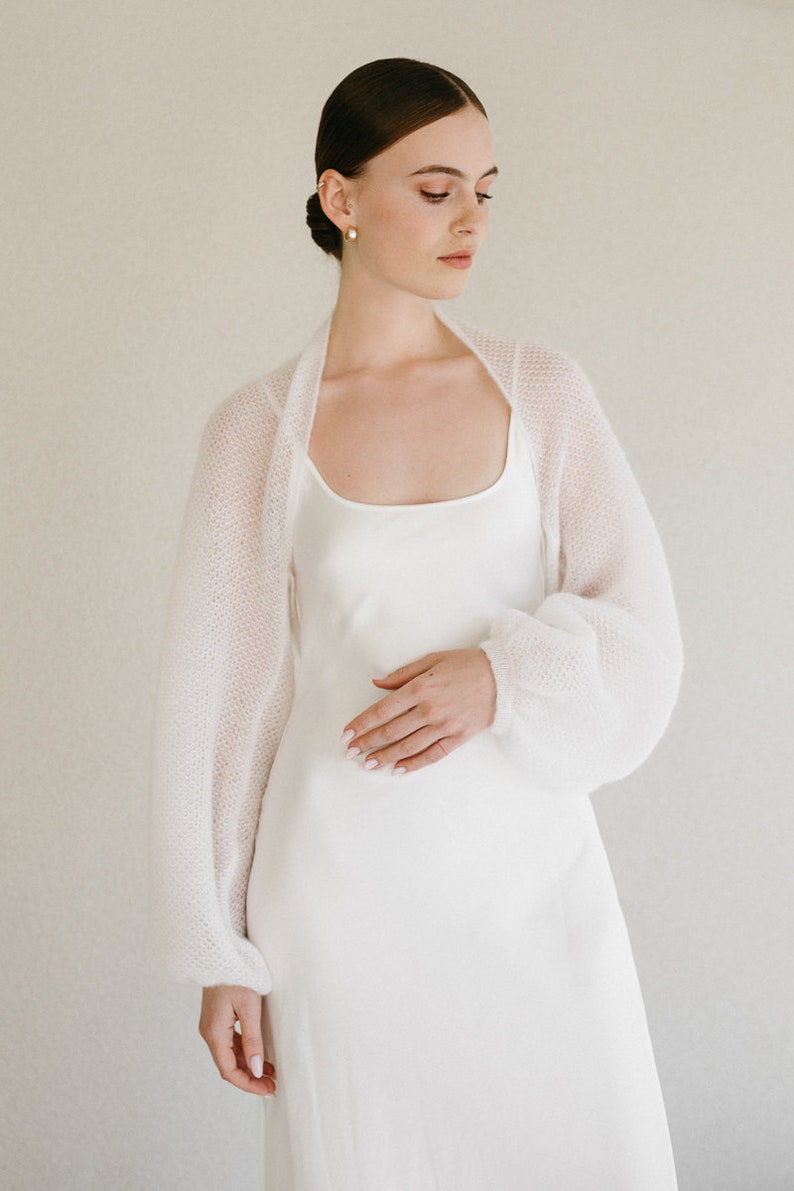 May include: A white knitted bolero with long, voluminous sleeves and a soft, textured finish, worn over a simple white slip dress with a scoop neckline. The garment features a lightweight, semi-sheer knit construction with ribbed cuffs.