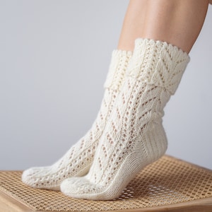May include: A pair of white knitted socks with a lacy pattern. The socks are folded down at the top and are resting on a wooden stool with a woven seat.
