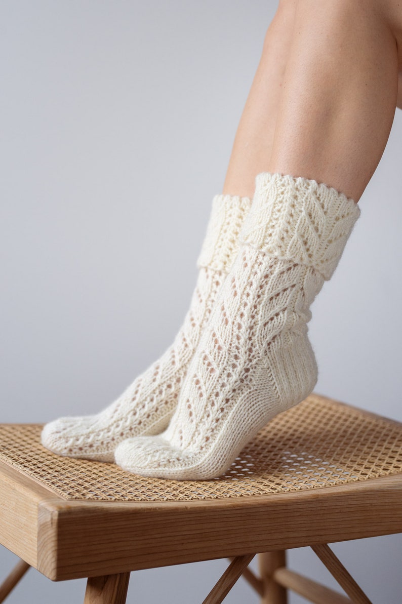 May include: A pair of white knitted socks with a lacy pattern. The socks are folded down at the top and are resting on a wooden stool with a woven seat.
